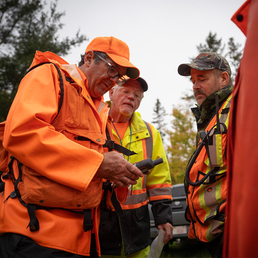 A group of search volunteers stand around someone holding a walkie talkie, watching carefully to their instruction.